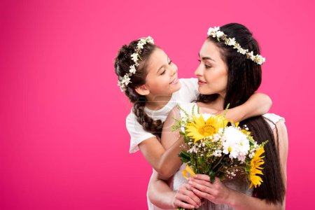 adorable happy daughter looking at beautiful mother holding flower bouquet isolated on pink