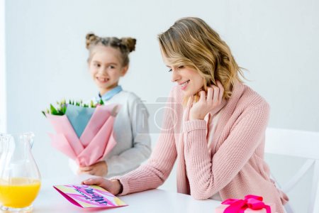 selective focus of happy mother looking at greeting postcard and daughter with bouquet of flowers standing near by, mothers day concept