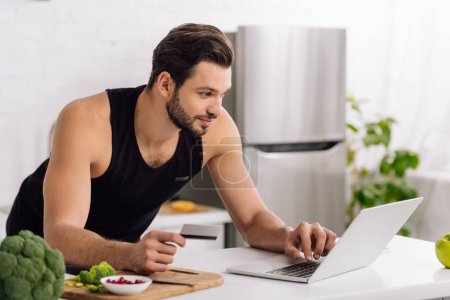 handsome man holding credit card while using laptop in kitchen 