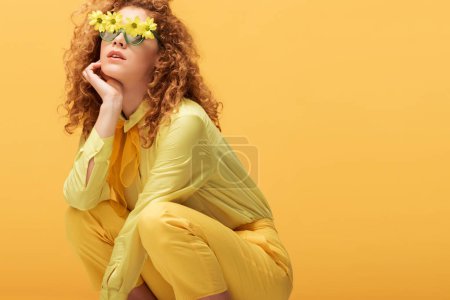 stylish redhead woman in sunglasses with flowers sitting isolated on yellow 