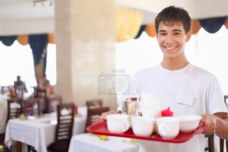 Young smiling affable waiter keeps tray with dishes at restauran