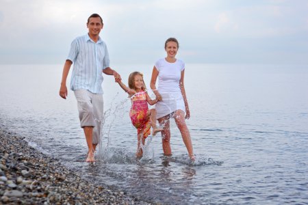 Happy family with little girl splashes feet water on beach in ev