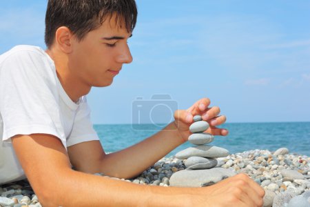 Teenager boy lying on stony seacoast, creates pyramid from pebbl