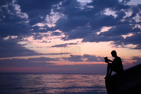 Silhouette guy sitting on breakwater in evening near sea, reads
