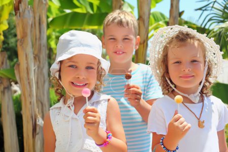 Smiling children three together eat lollipop in park