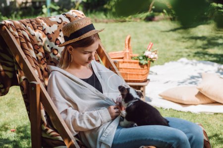 blonde girl in straw hat holding puppy while sitting in deck chair in garden during picnic