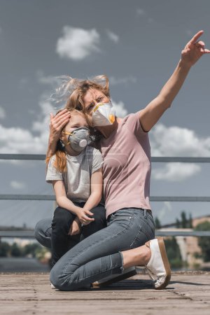 mother pointing on something to daughter in protective mask on bridge, air pollution concept