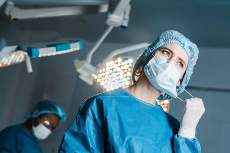 selective focus of nurse in uniform putting off medical mask in operating room