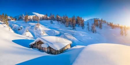 Panoramic view of beautiful winter mountain landscape with snow capped mountain cabin in the Alps in golden evening light at sunset