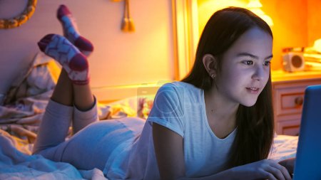 Portrait of beautiful girl lying on bed at night and using computer